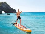 Άνδρας κάνει paddleboarding σε γαλαζοπράσινα νερά κοντά σε μια πέτρινη αψίδα.