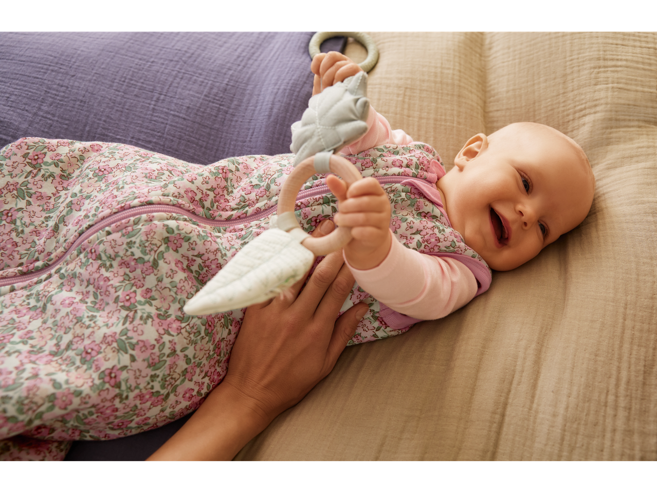 A baby in a floral sleep sack holding a wooden and fabric teether, smiling on a blanket.