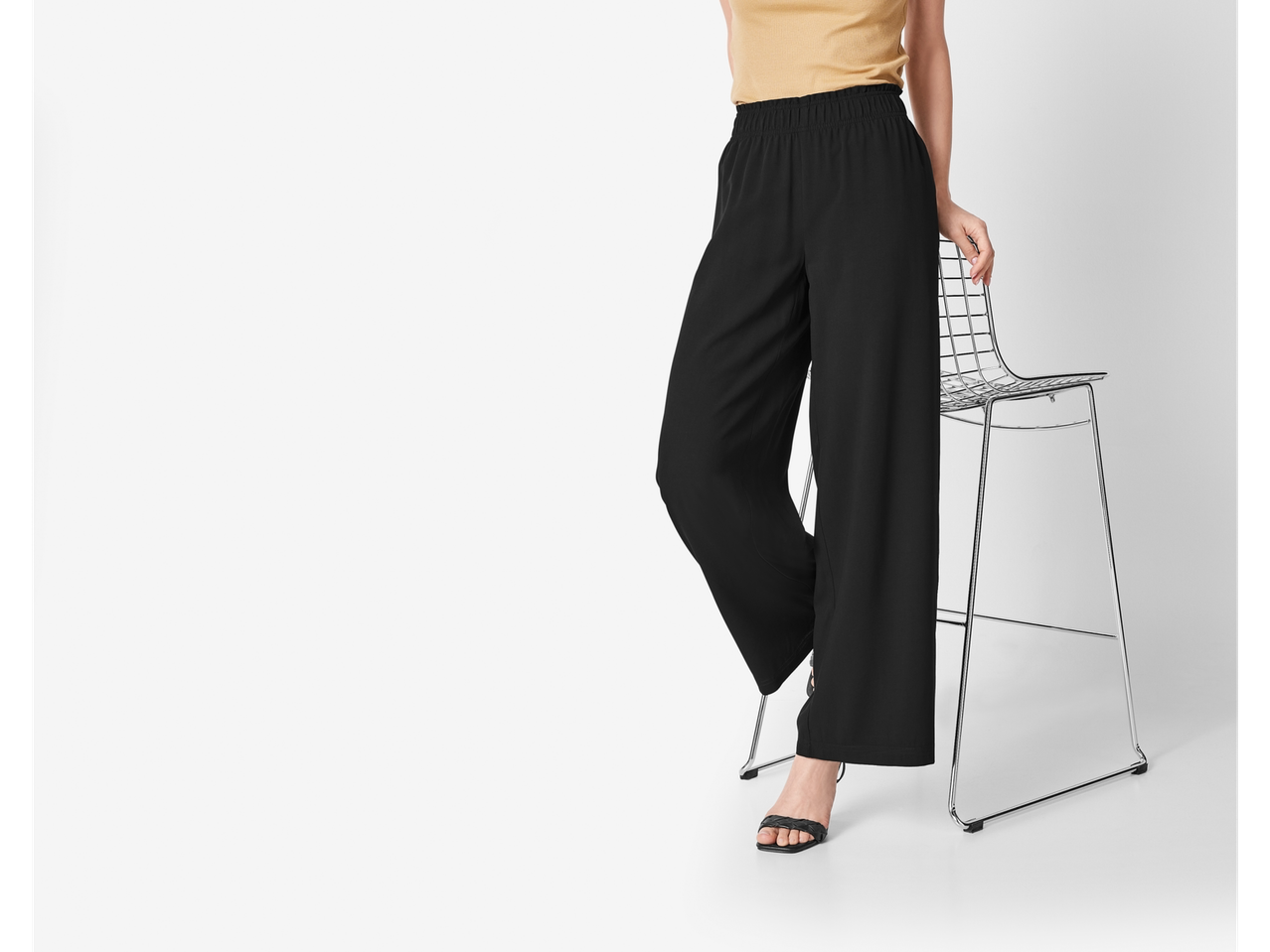 Pantaloni largi negri cu talie elastică, model pozând lângă un scaun metalic.