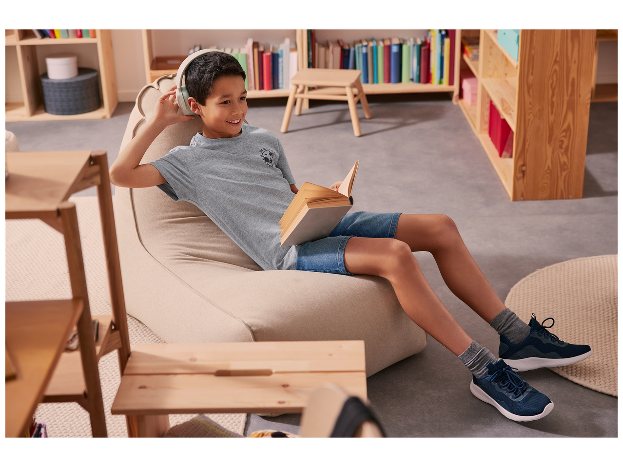 Un garçon lit un livre, assis sur un pouf, avec des écouteurs, dans une pièce avec des étagères.