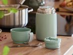 Light green food thermos, cup, and spoon on a wooden counter next to a pot of soup.