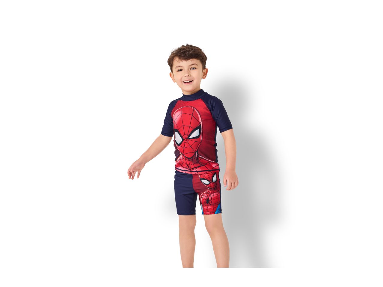 A young boy smiling and wearing a red and blue short-sleeved rash guard and swim shorts.