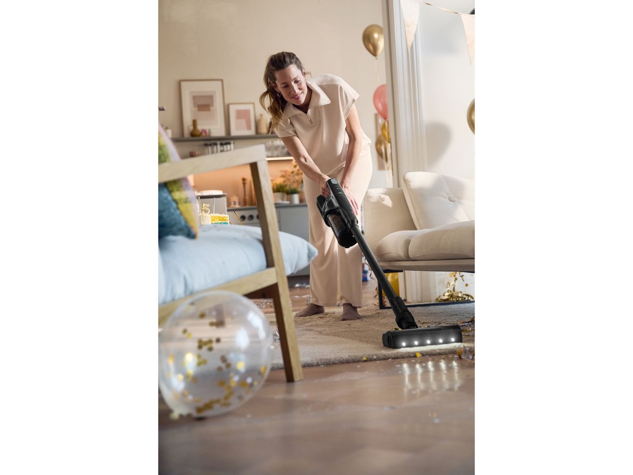 A woman uses a black cordless vacuum cleaner on a light wood floor.