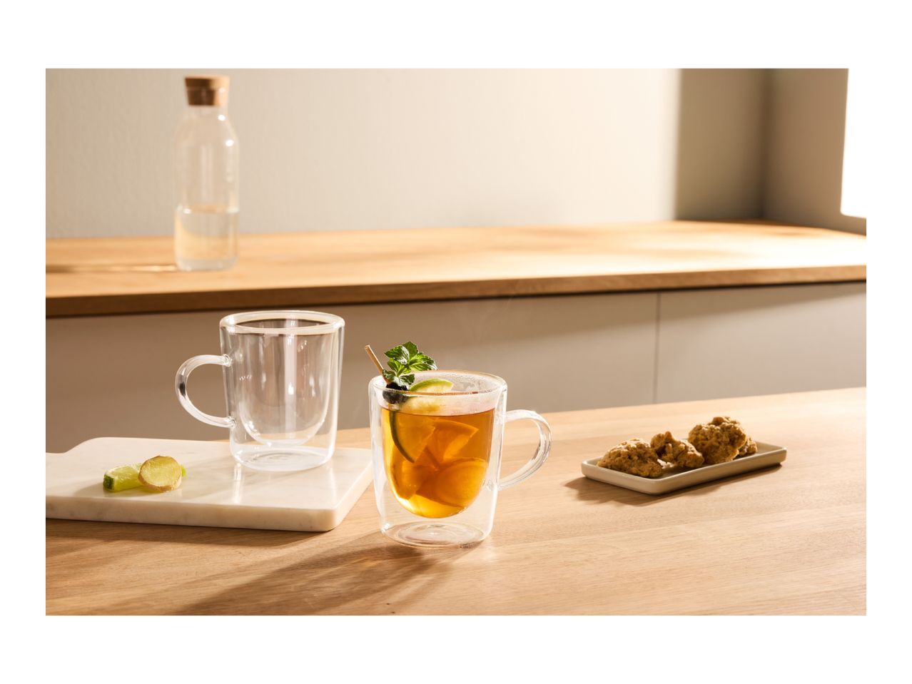 Two double-walled glass mugs, one with tea and garnishes, on a wooden kitchen counter.