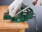 A person uses a file to shape a metal bar held in a green vise.