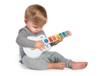 A toddler in a gray outfit sits on a white floor, playing a white and wooden Baby Einstein toy guitar.