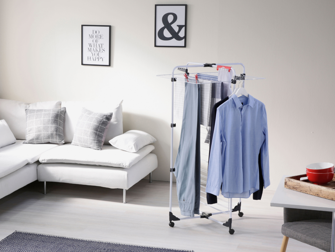 Сушилник за дрехи с колелца, на който висят ризи и панталони, в светла всекидневна.