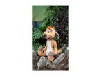 A plush meerkat toy with orange hair, sitting on a tree trunk amidst green and purple foliage.