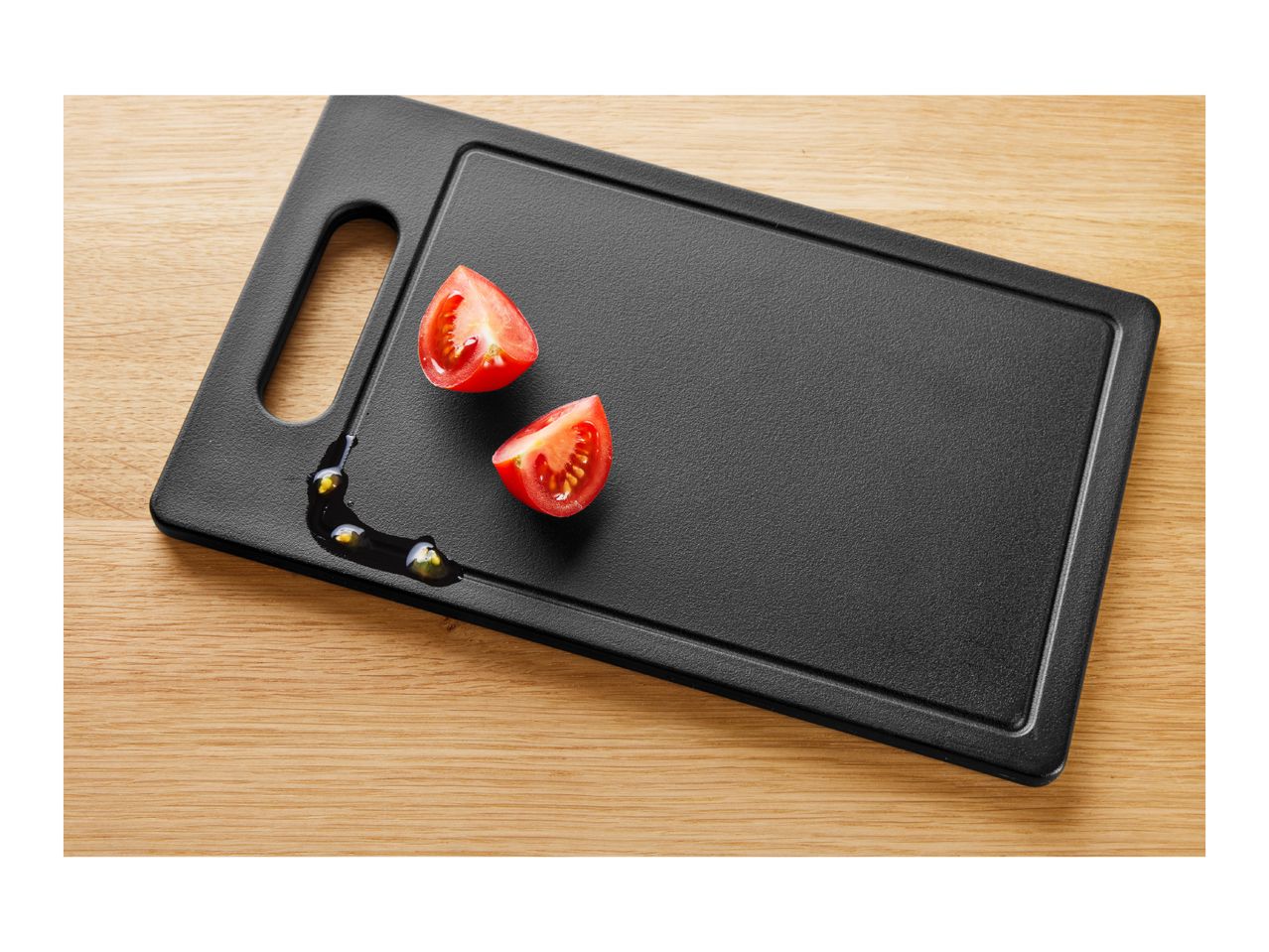Black cutting board with two tomato halves and olive oil on a wooden surface.