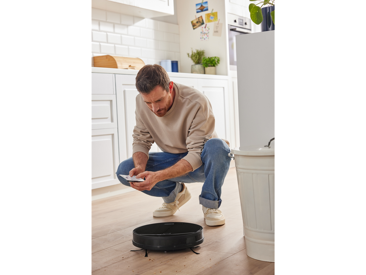 Homem controlando um aspirador robô Silvercrest com um smartphone em uma cozinha.