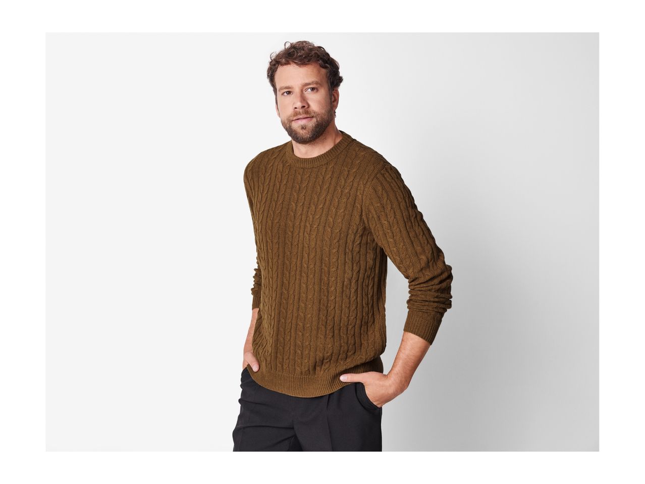 A man in a brown cable knit sweater and black pants, hands in pockets.
