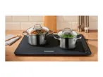Two stainless steel pots with glass lids on a black warming tray, filled with food, on a wooden kitchen counter.