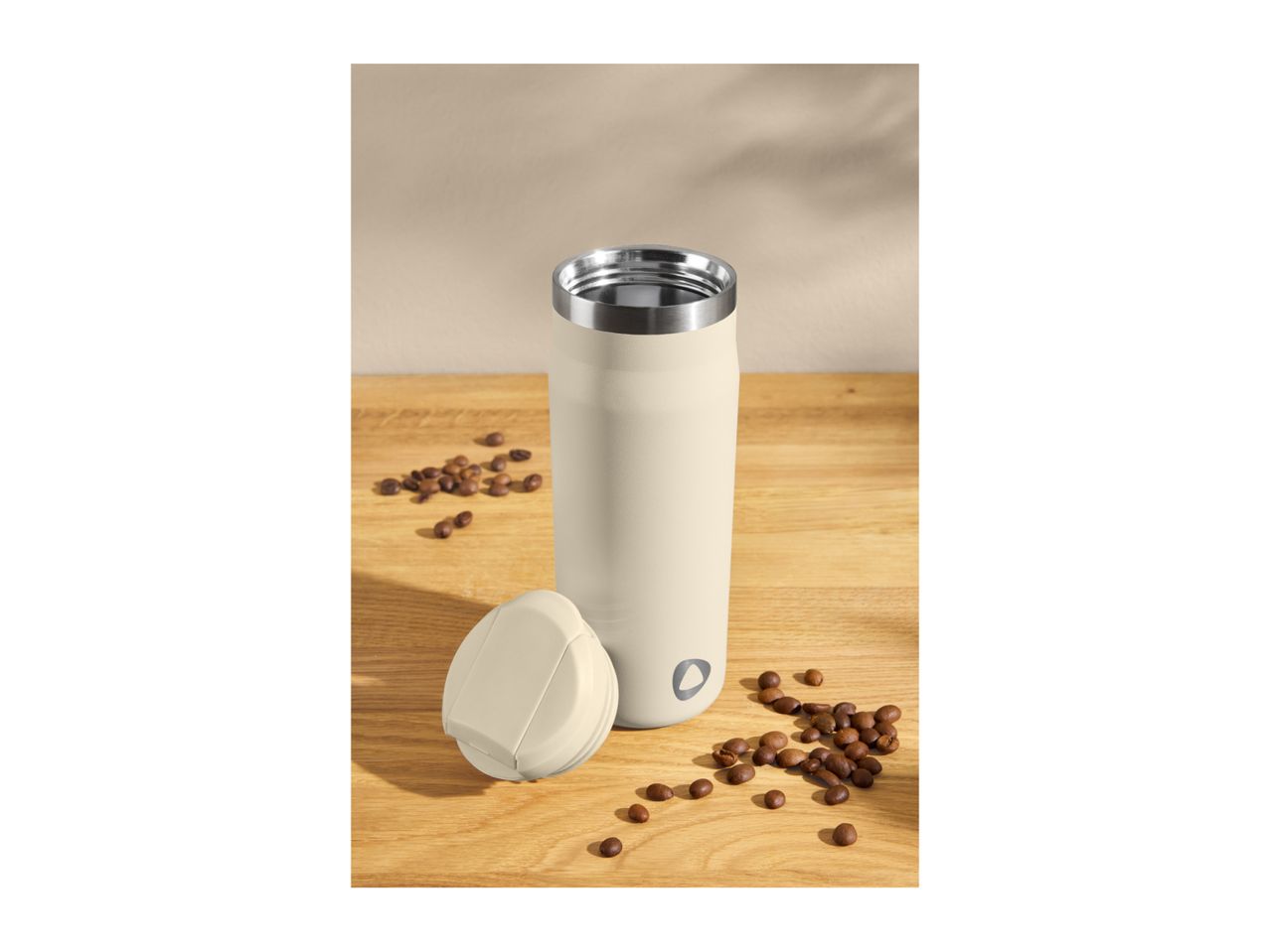 Cream-colored travel mug with lid and coffee beans on a wooden surface.