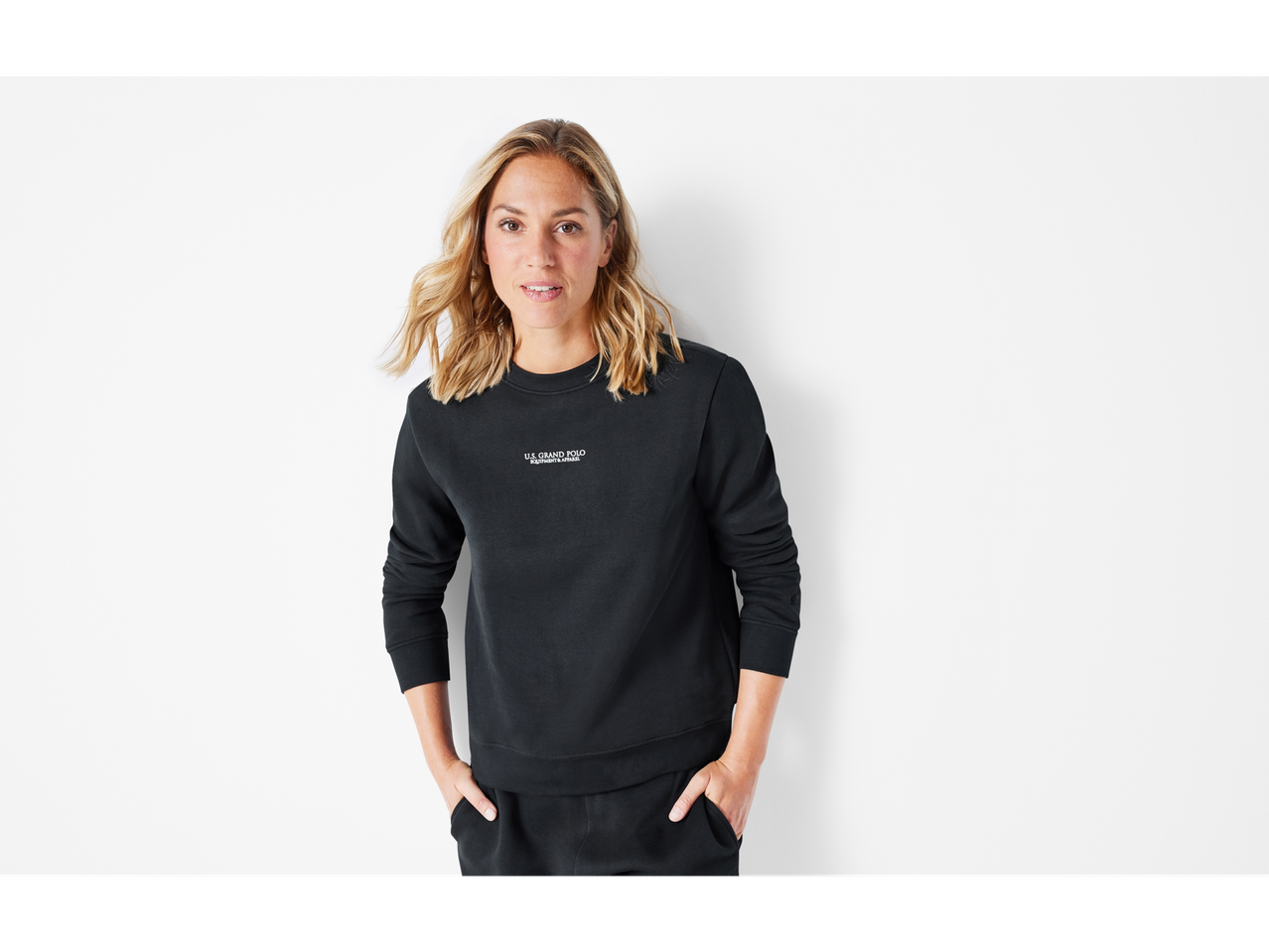 A woman in a black Esmara sweatshirt and sweatpants, hands in pockets, smiling.