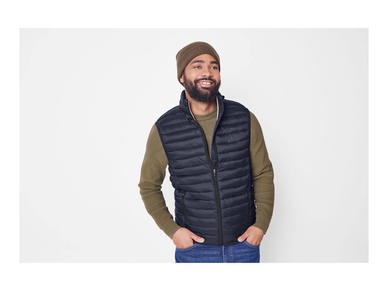 Man in a dark blue puffer vest, olive green sweater, and brown beanie smiling.