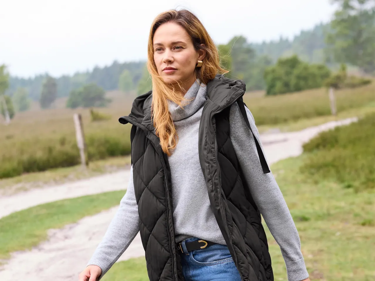 Egy nő szürke garbóban, fekete steppelt mellényben és farmerben áll egy ösvényen.