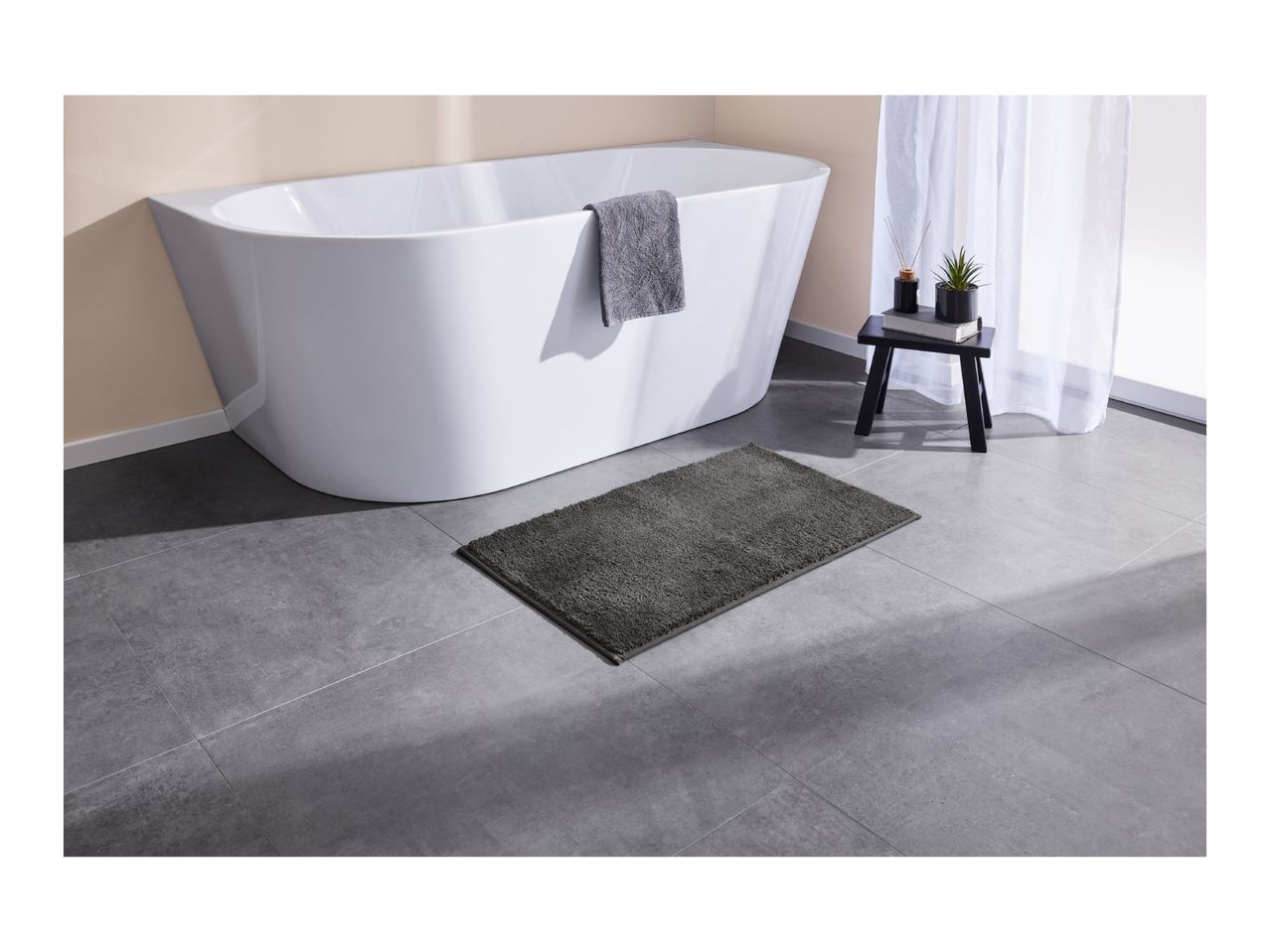 A white bathtub with a gray towel, a dark gray bath mat, and a small black table with decor.