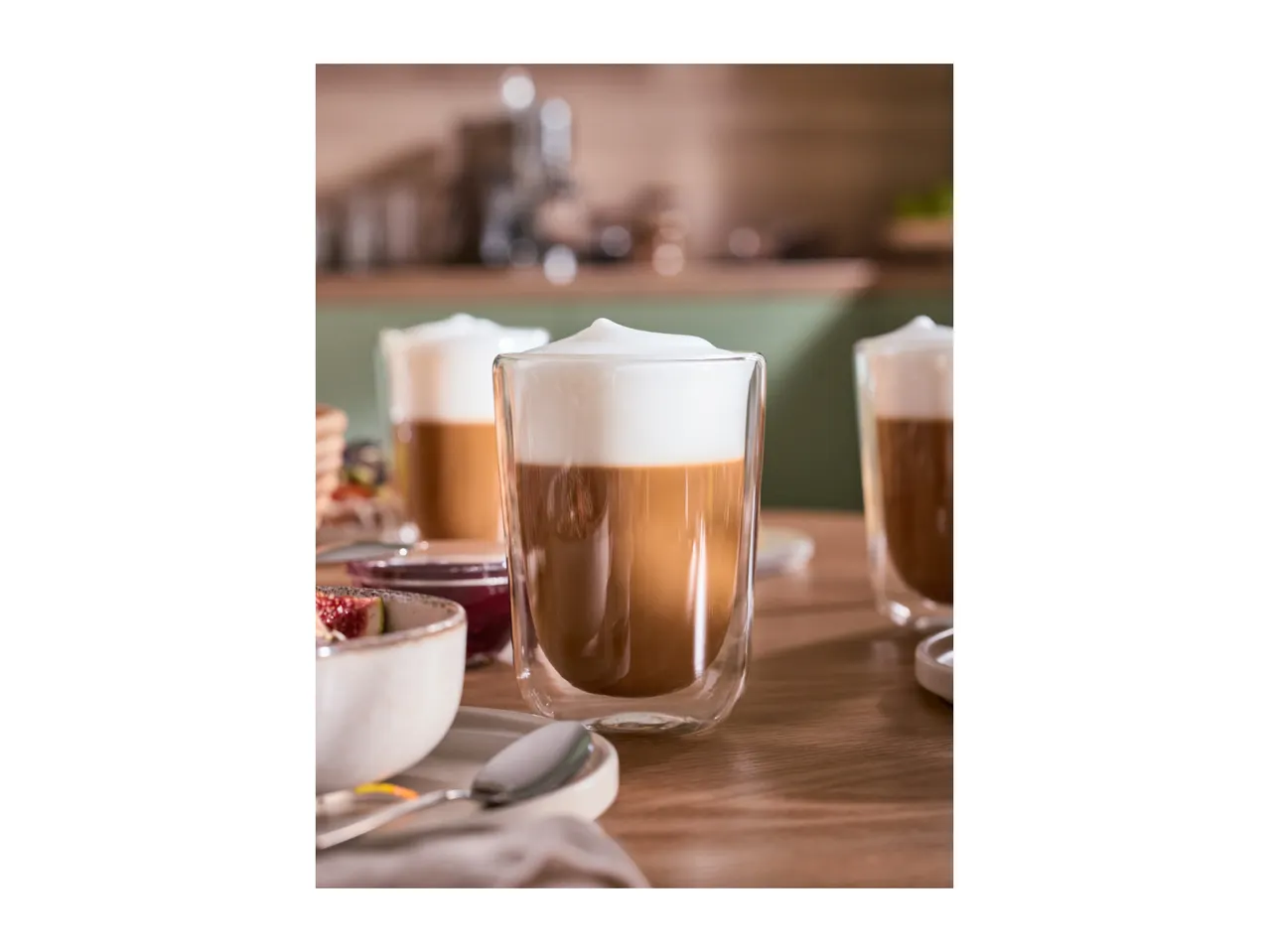 Two glasses of latte macchiato with foamed milk, a bowl of figs, and jam on a wooden table.