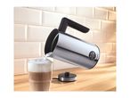 Stainless steel milk frother pouring foam into a glass of latte on a wooden counter.