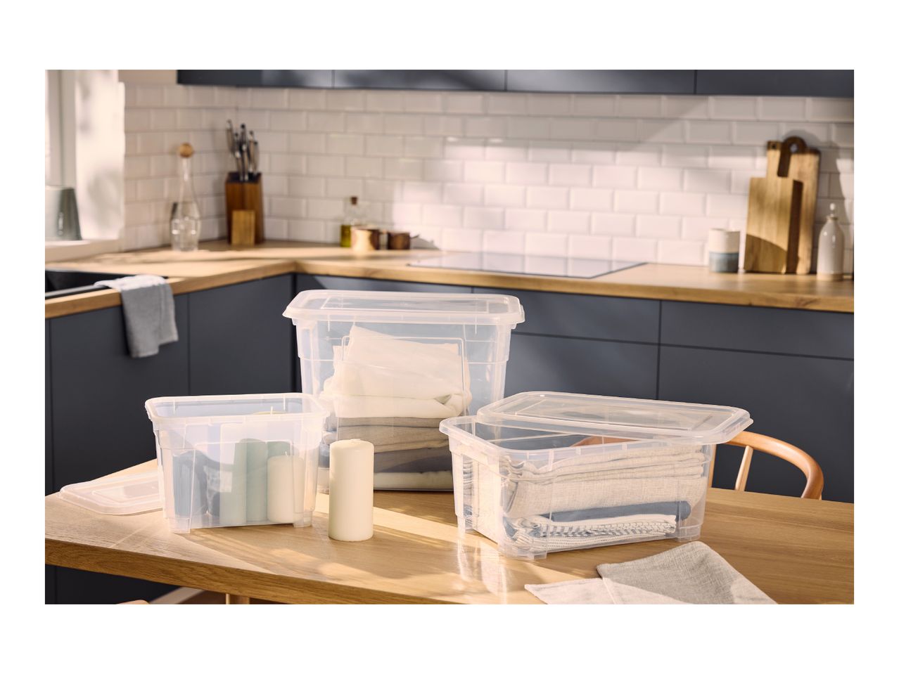 Three clear plastic storage boxes with lids on a wooden table in a kitchen setting.