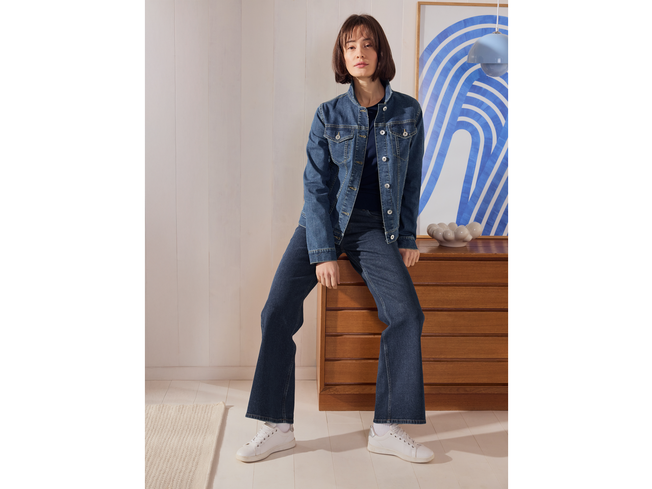 Mulher com camisola ESMARA e casaco de ganga, sentada numa cómoda de madeira.