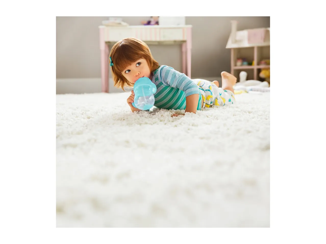 Toddler in pajamas drinking from a sippy cup while lying on a white rug.