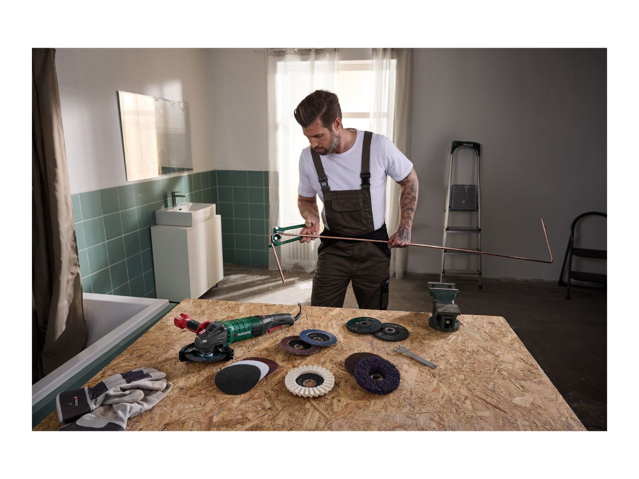 Man bending a copper pipe with a pipe bender, with an angle grinder and accessories on a workbench.