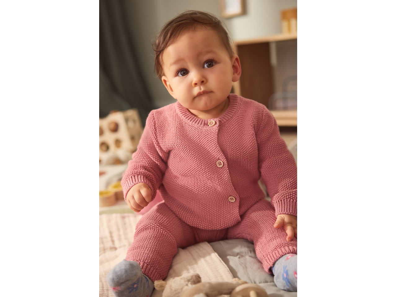 Baby in a pink knitted cardigan and trousers, sitting on a soft blanket.