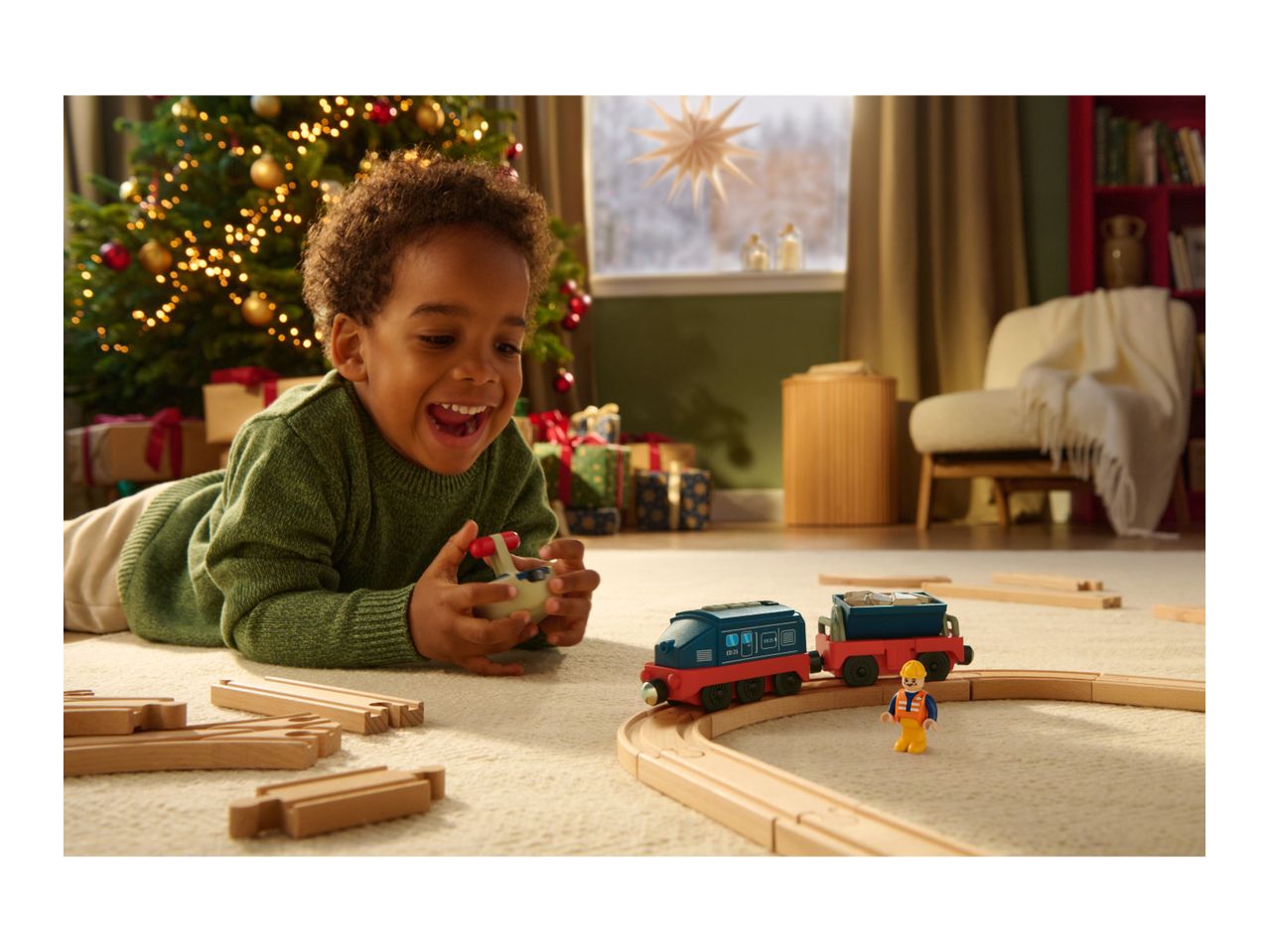 A happy child plays with a toy train set on a rug, with a Christmas tree in the background.