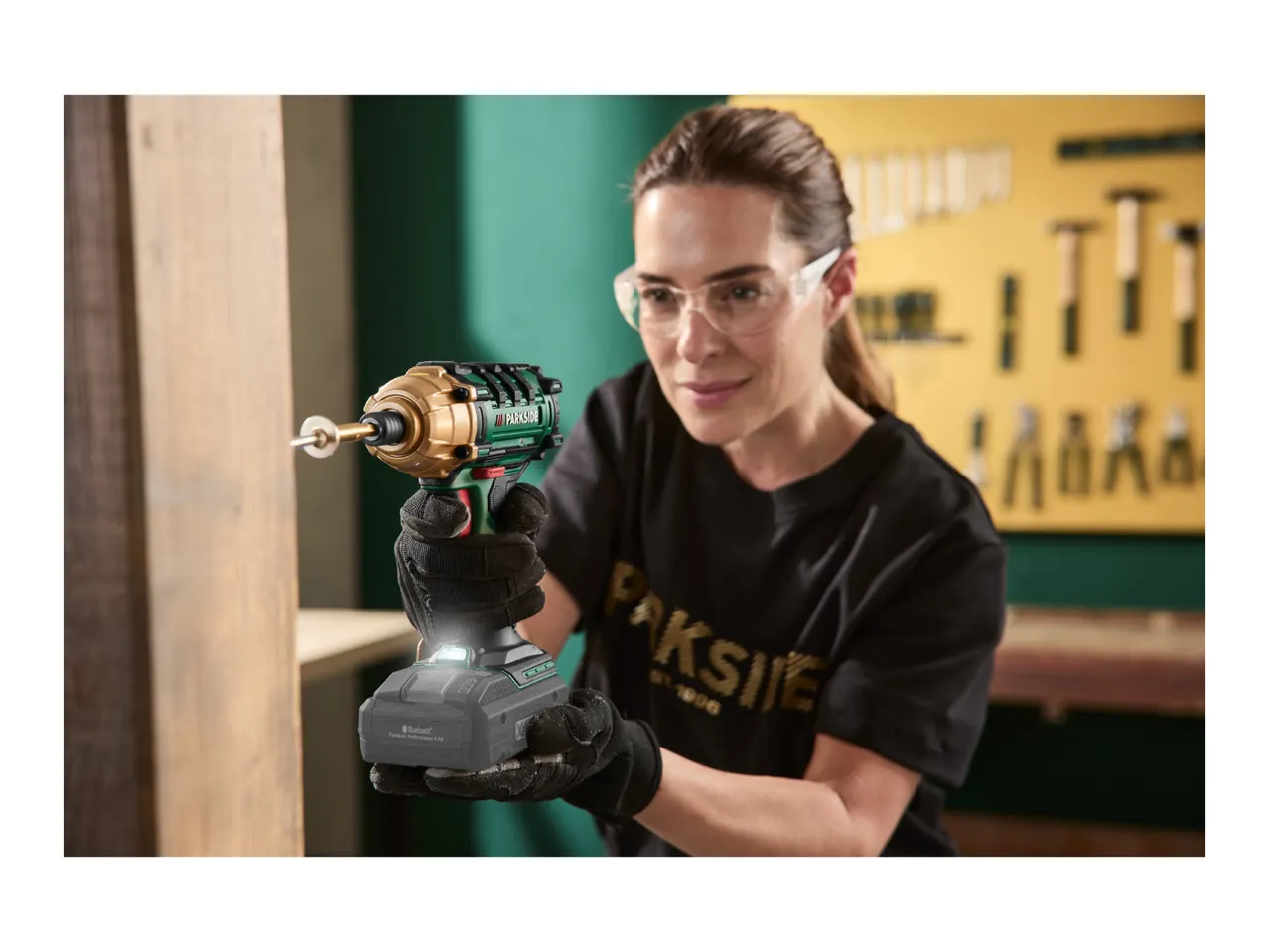 Woman in safety glasses using a cordless impact driver on wood in a workshop.