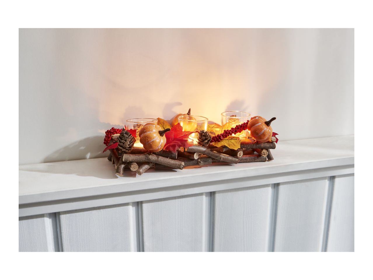 Autumnal candle holder with pumpkins, leaves, and berries on a white shelf.