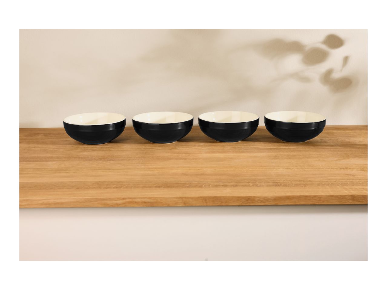 Four black and white bowls on a wooden surface with plant shadows on the wall.