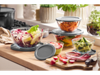 Saladas e vegetais picados em tigelas de vidro com tampas cinzentas numa bancada de cozinha.
