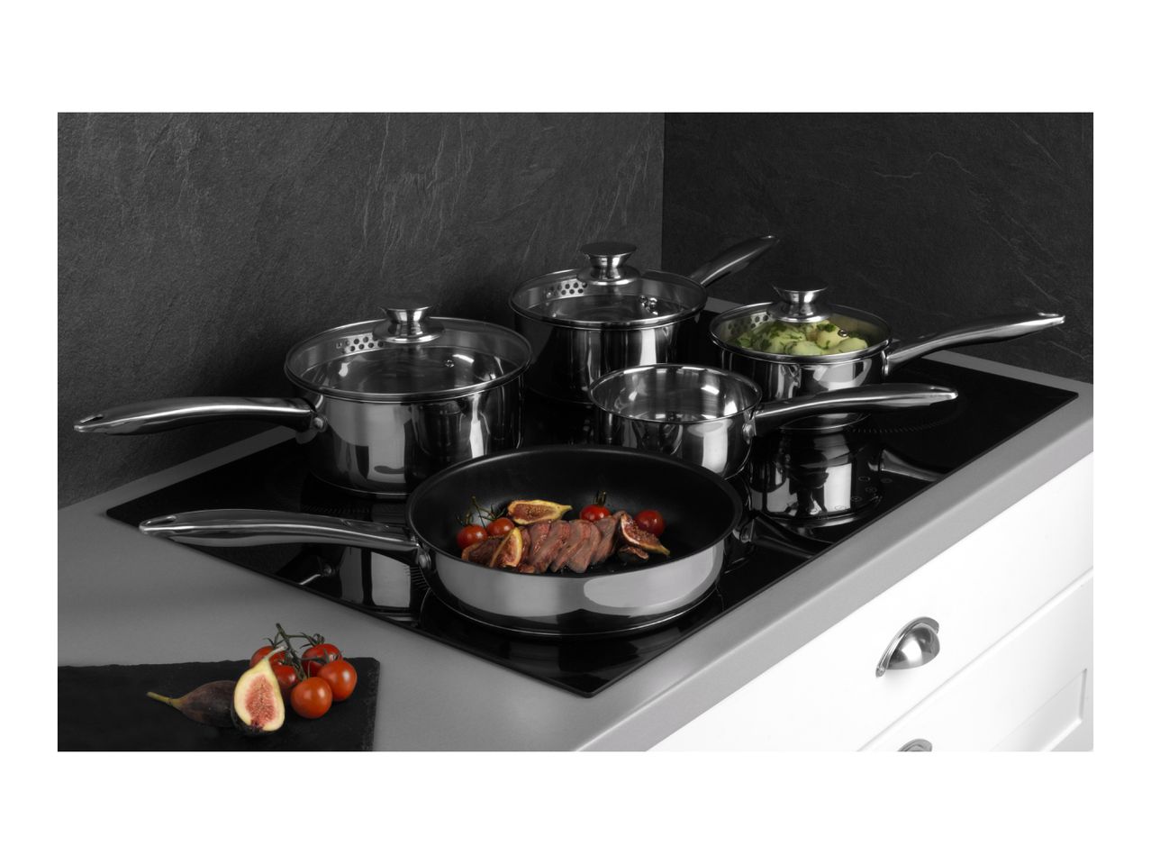 Stainless steel cookware set on an induction hob, with a frying pan cooking meat and figs.