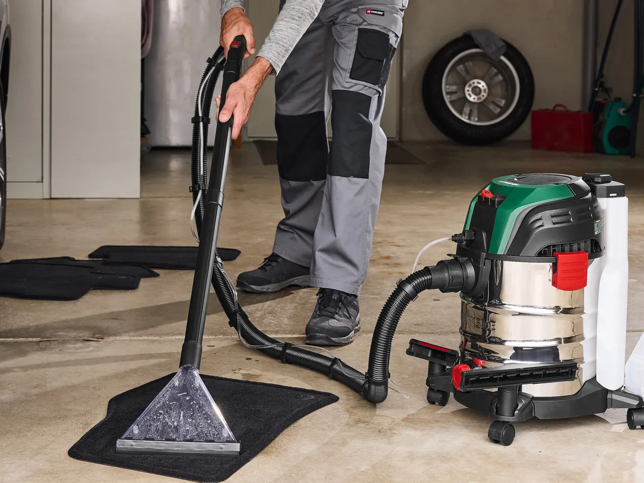A person using a wet/dry vacuum cleaner to clean a car mat in a garage.