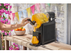 Aparat negru de granita turnând băutură portocalie într-un pahar pe o masă de lemn.