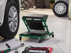 Green workshop creeper with tools and car battery charger next to a car wheel.