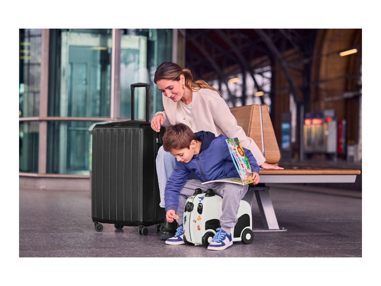 Woman and child with a black suitcase and a panda ride-on suitcase at a train station.