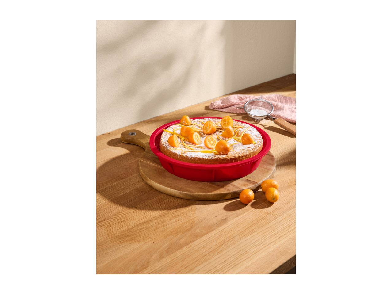 A kumquat cake in a red baking pan on a wooden board, with kumquats and powdered sugar.