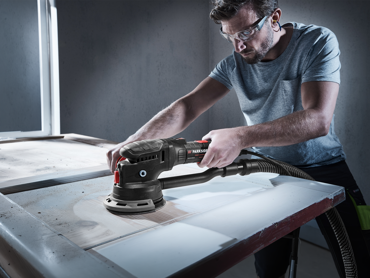 Άνδρας τρίβει μια ξύλινη επιφάνεια με τριβείο, φορώντας γυαλιά ασφαλείας και ωτοασπίδες.