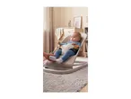Baby in a beige bouncer, with a hand touching their foot, in a neutral-toned room.