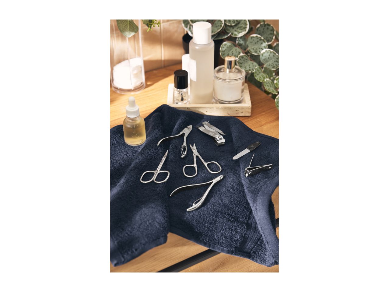 Manicure set with nail clippers, scissors, and file on a dark blue towel, with beauty products in the background.