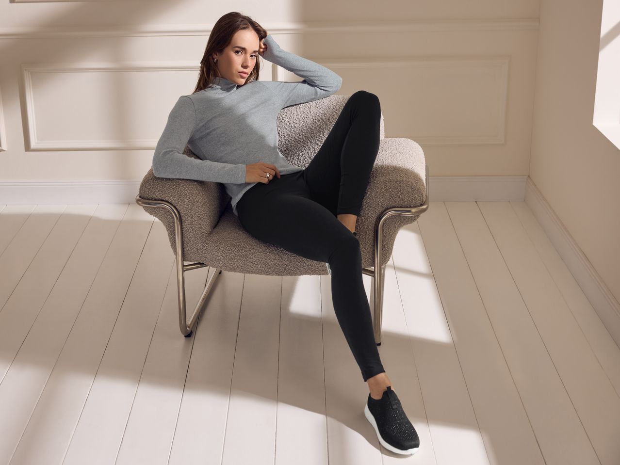 Woman in a grey turtleneck and black leggings sitting on a textured armchair.