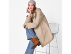 Woman in a beige raincoat, blue jeans, and brown boots, sitting on a metal chair.