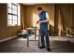 Bărbat în vestă și pantaloni de lucru Parkside® ține o bormașină la o masă de lucru.
