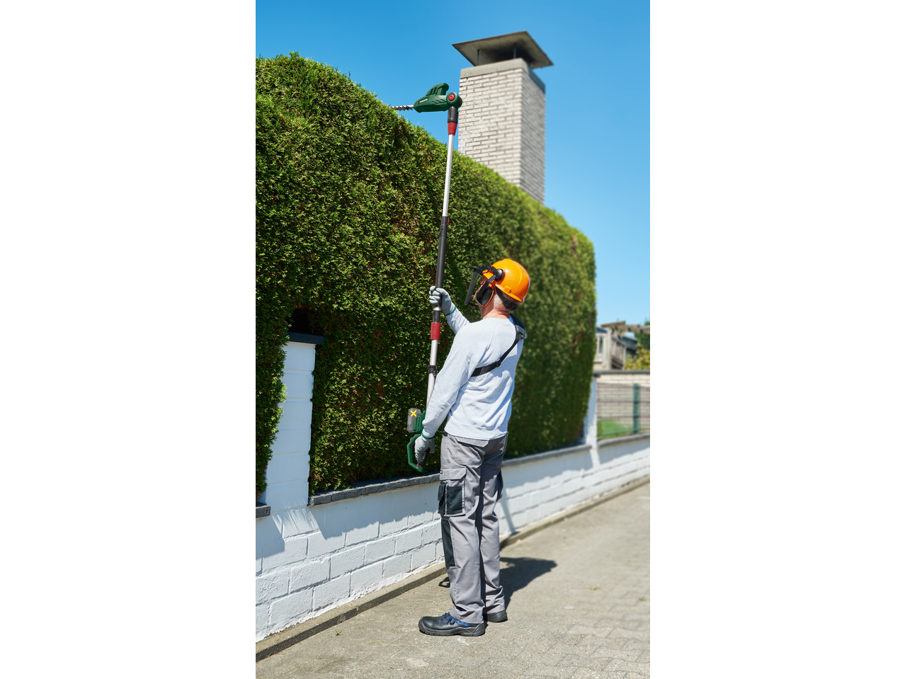 Man trimming a tall hedge with a long-reach hedge trimmer, wearing safety gear.