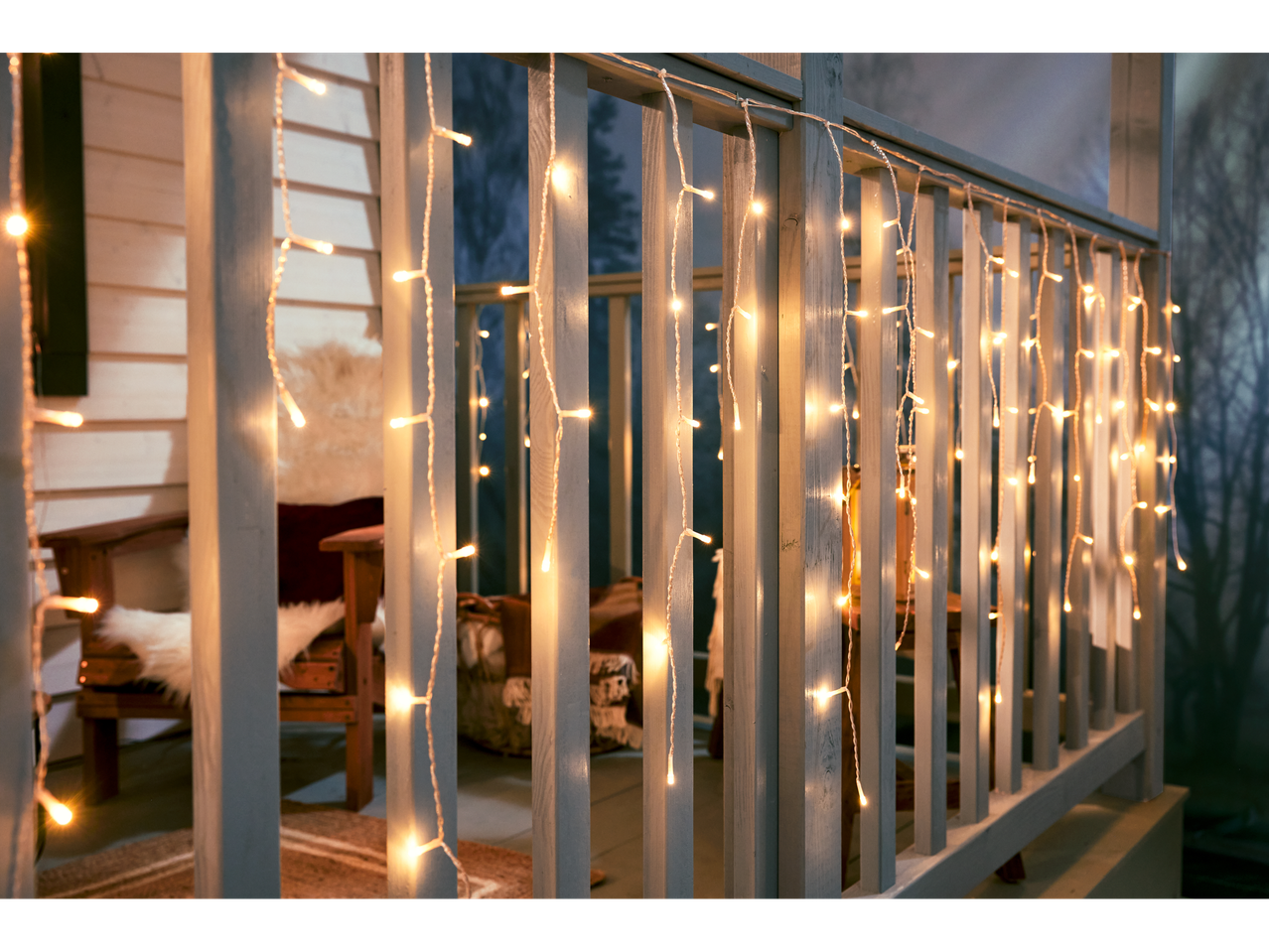 Porch railing with warm white icicle string lights, cozy outdoor seating in background.