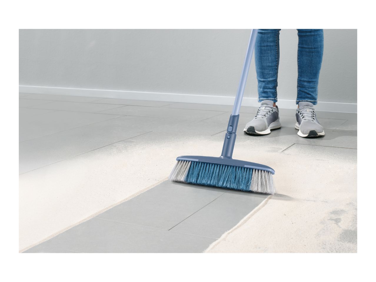 Person sweeping a light-colored floor with a blue and white broom, wearing jeans and sneakers.
