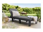 Dark grey sun lounger with cushions and a blanket on a patio, surrounded by greenery.