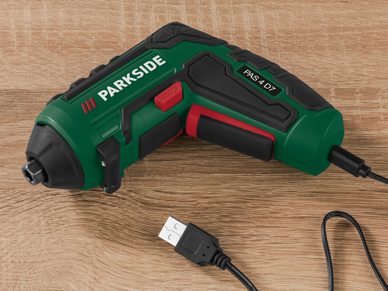 A Parkside PAS 4 D7 cordless screwdriver with USB charging cable lies on a wood surface.